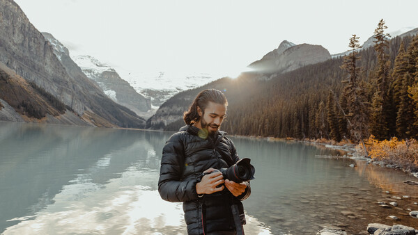 Photographer reviewing images on a camera beside a glacial lake surrounded by snow-capped mountains.