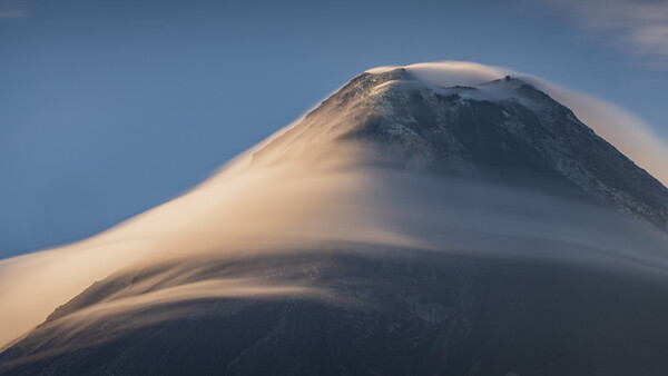Snow-capped volcanic cone rising through layered lenticular clouds at dawn.