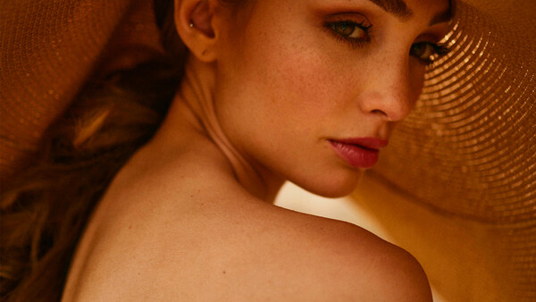 Woman's face and bare shoulder against warm, textured background with dramatic side lighting.