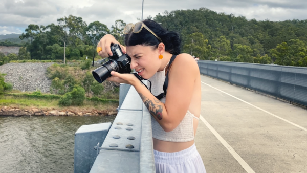 Photographer with camera on concrete bridge railing overlooking river and forested landscape.