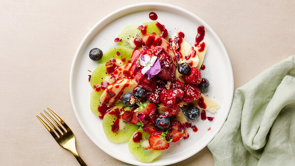 Overhead view of a plated fruit arrangement with kiwi slices, berries, and red fruit coulis on white ceramic plate.