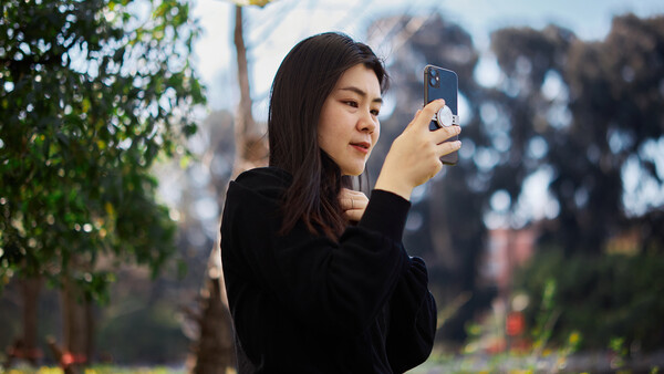 Woman holding smartphone horizontally to capture a photograph, with blurred urban landscape and green foliage in background.
