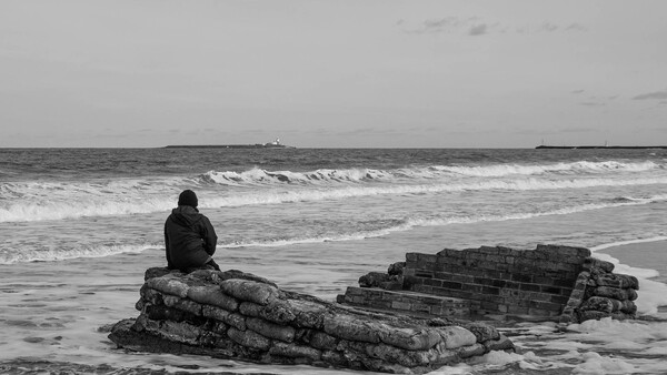 Person sitting on a textured rock formation in shallow ocean waves under an overcast sky.