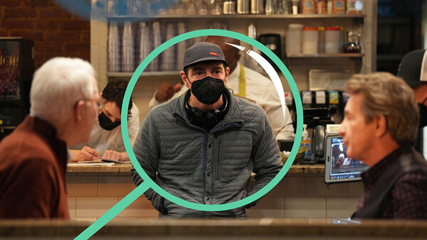 Man wearing a cowboy hat and black face mask stands behind a counter, highlighted with a green circle overlay.