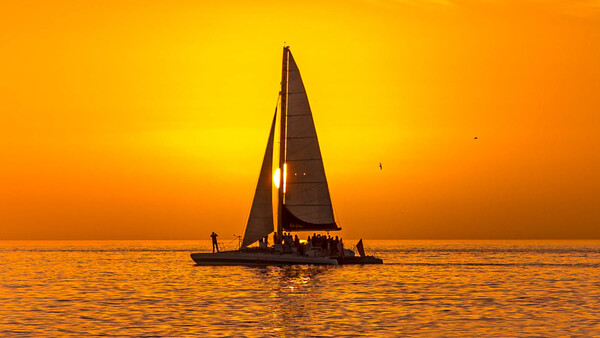 Silhouetted sailboat with passengers on calm ocean waters during golden sunset.