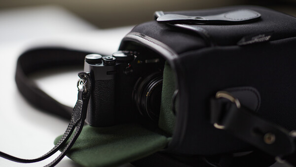 Compact rangefinder camera with black leather strap on olive green fabric bag.