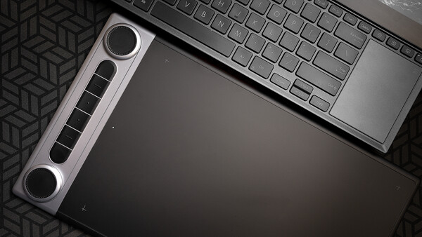 Flat lay of a graphics tablet with stylus pen positioned next to a laptop keyboard on a dark geometric patterned surface.