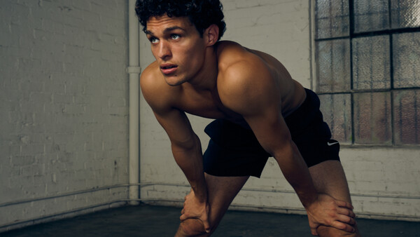 Shirtless man in athletic stance in industrial studio with concrete walls and barred window.