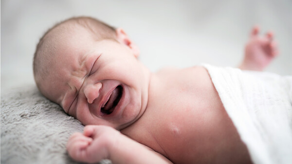Newborn baby yawning with eyes closed, wrapped in white fabric against a gray textured surface.