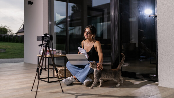 Woman with glasses crouching beside a cat while holding papers, with a camera on tripod and professional lighting setup on a modern patio.