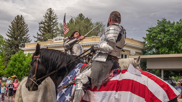 Two people in medieval armor on horseback with American flag draped across the horse.