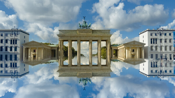 Classical European monument with tall columns reflected in a mirror-like sky effect, flanked by period buildings.
