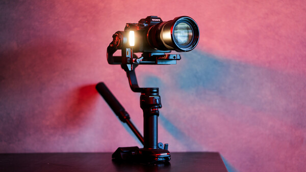 Camera mounted on a three-axis gimbal stabilizer with LED light attached, photographed against a pink and blue gradient background.