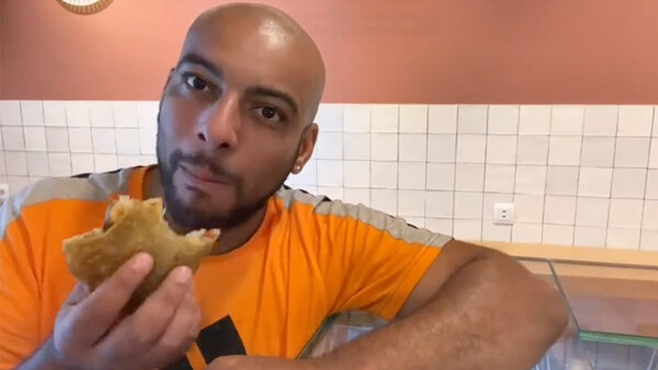 Man in orange shirt holding a sandwich in a tiled kitchen, looking at camera.