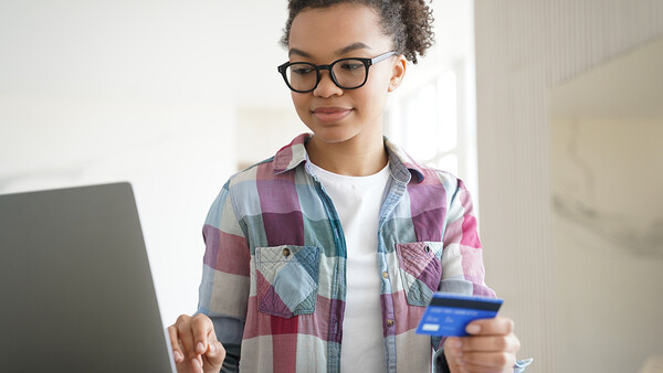 Young person wearing glasses and a plaid shirt holds a credit card while looking at a laptop screen.