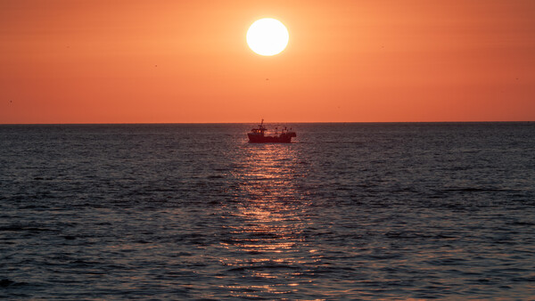 Silhouetted fishing vessel on calm water beneath a large sun during golden hour sunset.