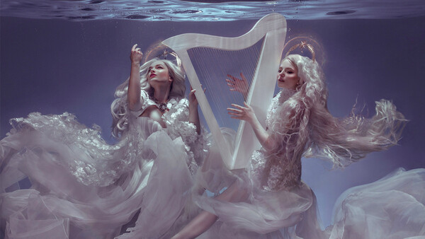 Two figures in flowing white gowns underwater with a harp, shot against purple-toned lighting.