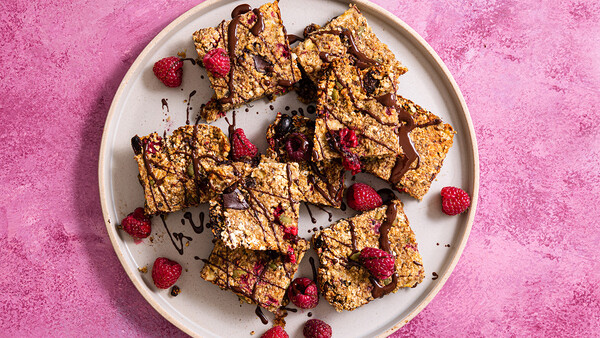 Golden granola bars with chocolate drizzle and fresh raspberries on a white plate against a pink textured background.