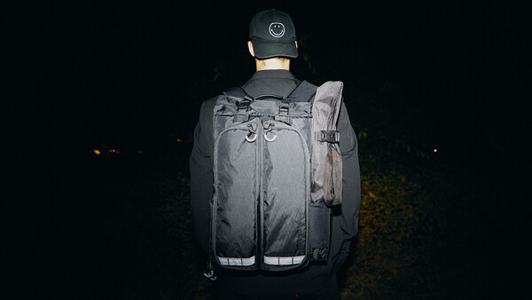 Person wearing a gray backpack and cap photographed from behind at night against a dark background.