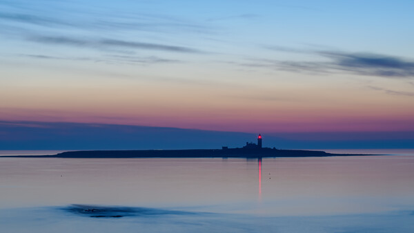 Lighthouse beam reflects across still water at twilight, with distant forested shoreline silhouetted against purple and blue sky.