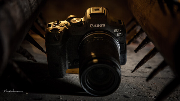Canon EOS R7 mirrorless camera with attached lens on textured surface with dramatic side lighting.