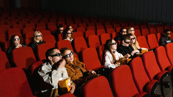 Audience members seated in a movie theater with red seats, some wearing glasses and holding popcorn.