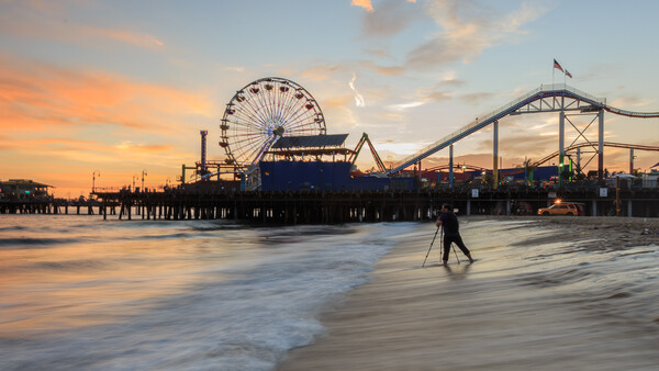 Surfer walking into the ocean at sunset with an amusement park pier and roller coaster in the background.