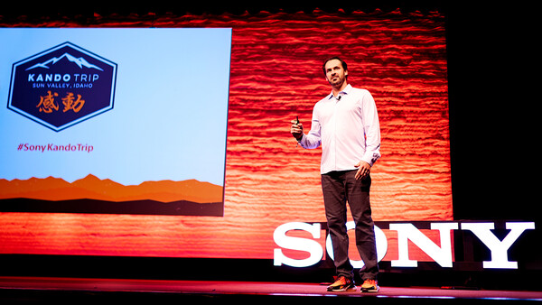 Speaker presenting on stage at a Sony event with red brick backdrop and illuminated signage.
