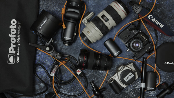 Professional photography gear laid flat on a textured dark surface, including camera bodies, lenses, lighting equipment, and cables with orange straps.
