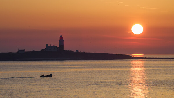 Silhouetted lighthouse on a peninsula at sunset with a small fishing boat in the foreground water.