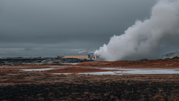 Geothermal power plant with massive white steam plumes rising from industrial structures across barren volcanic landscape.