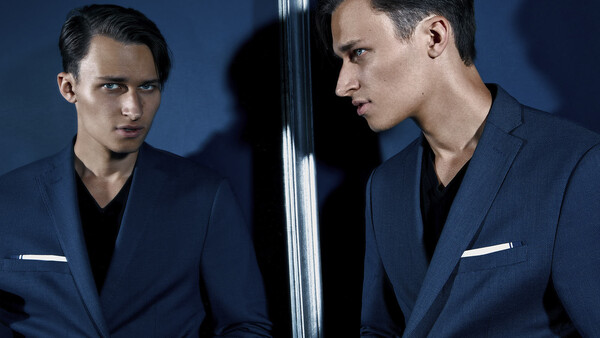Man in navy blazer looking at his mirror reflection against blue background.