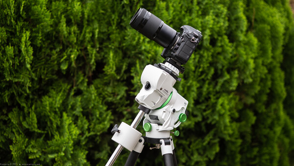 White and green motorized camera pan-tilt head mounted on tripod with DSLR camera and telephoto lens against green foliage background.