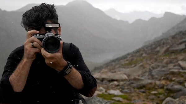 Photographer holding a DSLR camera up to his face while standing in a mountainous landscape during overcast conditions.