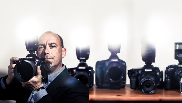 Photographer holding a Nikon DSLR camera with flash, surrounded by professional camera bodies on a wooden surface.