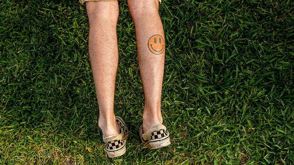 Person standing on grass wearing khaki shorts, checkered sandals, and an orange smiley face tattoo on their calf.