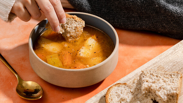 Hand dipping whole grain bread into a bowl of golden soup garnished with black pepper.