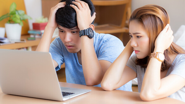 Two young adults looking at a laptop screen with concerned expressions while sitting at a wooden desk.