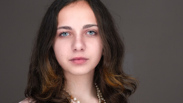 Portrait of a young woman with long dark hair and blue eyes against a neutral gray background.