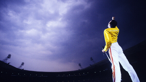 Performer on stage in yellow and white outfit against dramatic purple twilight sky and stadium lighting.