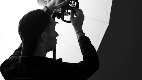 Silhouetted photographer holding a camera to their face while adjusting settings, shown in profile against a white backdrop.
