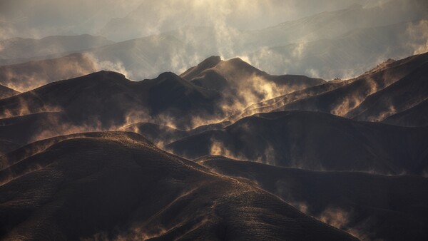 Wildfire smoke billows across mountainous terrain at golden hour, silhouetting ridgelines against dramatic clouds.