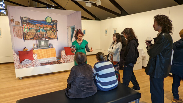 Gallery visitors observe an interactive bedroom display installation with a woman presenter in a green shirt explaining the exhibit.