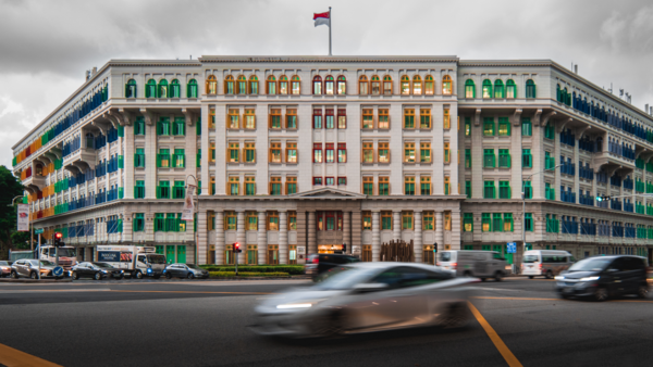 Colorful corner building with green and yellow-gold window frames and a flag on top, with blurred vehicles passing in foreground.