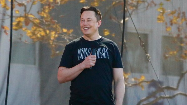 Man in black t-shirt speaking with microphone against backdrop of golden autumn foliage.