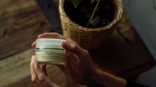Hand holding a cosmetic jar labeled Lesina Black Star Moisturizer against a wooden surface with woven basket backdrop.