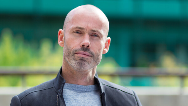 Bald man with facial hair wearing a dark jacket over a gray shirt, photographed outdoors with blurred green background.