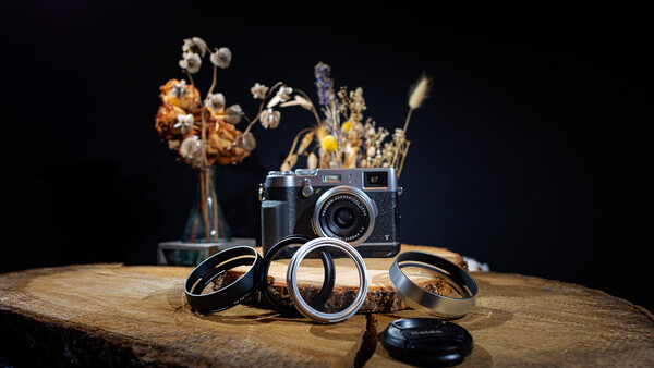 Rangefinder camera with brass lens rings and dried flowers arranged on weathered wood against black background.