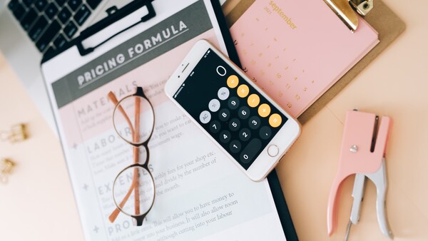 Flat lay workspace with laptop, smartphone showing calculator app, eyeglasses, pink notebook, and printed documents on beige surface.