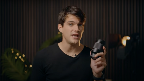 Man in black v-neck shirt holding a microphone while speaking, shot against a dark corrugated backdrop.